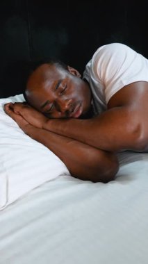 Afro-Amerikalı bir adam, sabah güneşiyle çevrili, rahat bir otel yatağında yavaşça uyanıyor. Huzurlu atmosfer, güne rahat bir ortamda huzurlu bir şekilde başlamanın altını çiziyor.