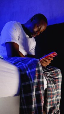 Pijamalı Afro-Amerikalı bir adam geceleri yatağında uykusuzlukla boğuşuyor ve telefonunu kontrol ediyor. Loş, mavi tonlu ışıklandırma onun huzursuz durumunu vurguluyor.