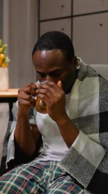 Sıcak bir battaniyeye sarılmış, soğuk algınlığını yatıştırmak için sıcak çay içen Afro-Amerikalı bir adam. Etrafı ilaçlarla çevrili halde, iyileşme sürecinde sıcaklık ve rahatlık arıyor.