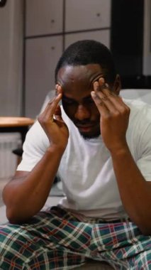 Kanepede oturan bir adam, acı dolu bir ifadeyle şakaklarını bastırıyor, baş ağrısı ve duygusal sıkıntı belirtileri gösteriyor, muhtemelen depresyon ya da stres.