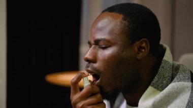 Ağrıyı ve rahatsızlığı azaltmak için boğazını spreyleyen bir adamın yakın çekimi. Sıcak bir battaniyeye sarılmış halde, sıcak bir iç mekanda soğuk semptomların tesellisini buluyor.