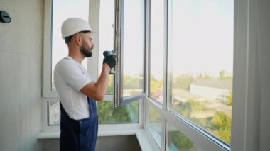 Deneyimli inşaat işçisi, yüksek katlı bir apartmana güvenli ve enerji tasarruflu bir mühür takmak için bir pencere açmak için elektrikli matkap kullanıyor.