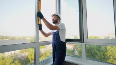 Başlık ve eldiven takan yetenekli inşaat işçisi, montaj sırasında pencere hizasını kontrol etmek, hassasiyet ve istikrar üzerine yoğunlaşmak için ruh seviyesini kullanır.