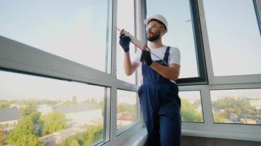Profesyonel inşaat işçisi pencere kenarlarına köpük macunu sürüyor, yalıtımı artırıyor ve modern yüksek katlı bir apartmanda hava sızıntılarını önlüyor