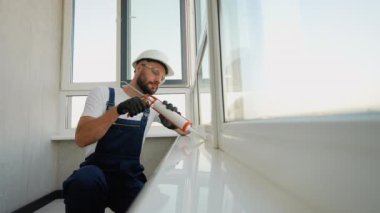 Koruyucu ekipmanlı inşaat işçisi, modern bir dairede hava geçirmez yalıtım ve enerji verimliliğini sağlamak için pencere çerçevesini sabitlemek için köpük dolgu maddesi kullanıyor.