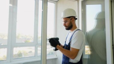 Koruyucu donanımlı yetenekli inşaat işçisi, modern yüksek katlı bir daireye güvenli ve hassas bir şekilde uyması için pencere açmak için elektrikli matkap kullanıyor.
