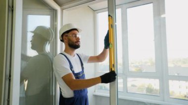 Güvenlik donanımlı profesyonel inşaat işçisi, modern bir binada güvenli bir kurulum için doğruluk ve istikrar sağlayan bir pencere çerçevesini hizalamak için ruh seviyesini kullanır.