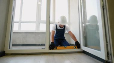 Koruyucu giysiler içinde yetenekli bir inşaat işçisi, bir kapı çerçevesini hizalamak için bir ruh seviyesini dikkatlice kullanır, hassasiyet, istikrar ve modern bir daireye güvenli bir uyum sağlar.