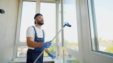 Eldivenler ve koruyucu giysiler içinde kendini işine adamış bir işçi, modern bir apartman ortamında tertemiz ve çıplak bir son garantilemek için büyük bir pencereyi temizlemek için bir cam sileceği kullanıyor.