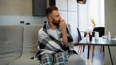 Sıcak ekoseli battaniyeye sarılı hasta bir adam soğuk belirtilerini gidermek için burun spreyi kullanır. Etrafı ilaçlarla çevrili ve rahatlamak için çay içer.