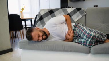Ekose pijamalı bir adam, karnını yoğun bir ağrı içinde kanepeye uzanmış, muhtemelen şiddetli bir mide ağrısı ya da apandisit sorununa işaret ediyor.