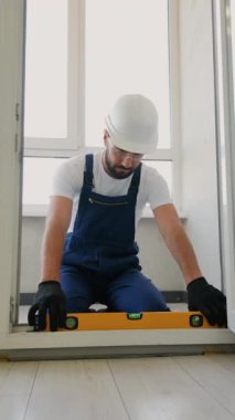 Koruyucu giysiler içinde yetenekli bir inşaat işçisi, bir kapı çerçevesini hizalamak için bir ruh seviyesini dikkatlice kullanır, hassasiyet, istikrar ve modern bir daireye güvenli bir uyum sağlar.