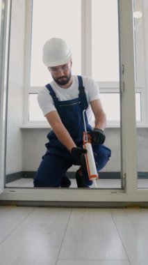 Güvenlik donanımlı profesyonel inşaat işçisi, caulking tabancasıyla pencere çerçevesine dolgu macunu sürüyor, hava geçirmez yalıtım sağlıyor ve modern bir daireye güvenli bir şekilde sığıyor.