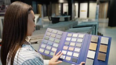 Bir kadın, sergi salonunda bir palet renk örneklerini karıştırarak ev dekorasyonu ve tasarım ihtiyaçları için mükemmel bir eşleşme bulma seçeneklerini dikkatle düşünür.