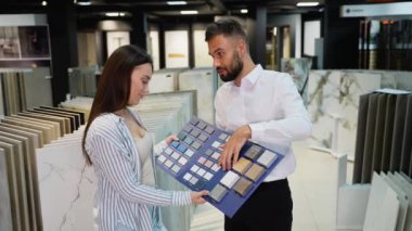 Satış Danışmanı Tay Seçimi ile Kadın Müşteriye Yardım Ediyor. Bir satıcı, sergi salonundaki çeşitli renk ve doku seçeneklerini göstererek kadın müşteriye fayans seçiminde yardımcı olur.