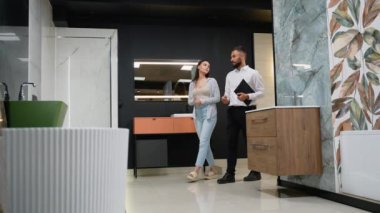 Bir satış danışmanı müşteriye fayans galerisindeki banyo dekorasyonu seçeneklerinde rehberlik eder, banyo tasarımını geliştirmek için şık fayanslar ve fikstür kombinasyonlarını vurgular.
