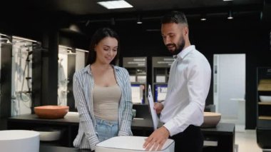 Bir satış danışmanı, müşteriye bir su tesisatı galerisinde modern bir lavabo tasarımı gösterir, özelliklerini açıklar ve banyo ya da mutfak projesi için ideal fikstürü seçmesine yardım eder.