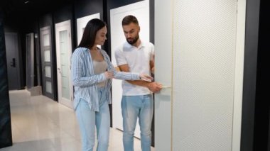 Bir çift, sergi salonunda kapı seçeneklerini inceleyerek evlerinin tasarımı ve işlevselliğini tartışıyor. Tadilatları için stil, renk ve kaliteyi düşünüyorlar.