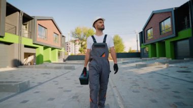 Çalışan bir müteahhit ve sert bir şapka, modern yeşil ve tuğla bir kulübenin yanında yürürken bir alet çantası taşıyor, profesyonelliği gösteriyor ve projeye odaklanıyor