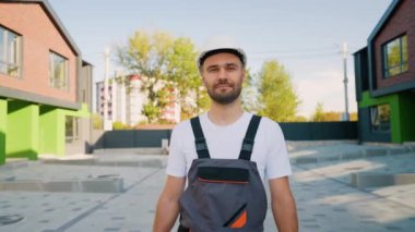 Sert şapkalı ve üniformalı bir inşaat işçisi, modern yeşil ve tuğladan bir kulübenin yanında duruyor. Profesyonelliğini ve çalışmaya hazır olduğunu gösteriyor.