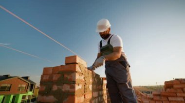 Hintli inşaat işçisi yeni bir ev için ustalıkla tuğla duvar örüyor, modern bir inşaat alanında güneşli gökyüzünün altına havan topu sıkıyor.