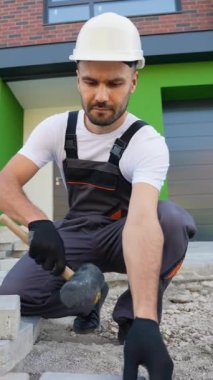 Sert bir şapka ve eldivenle inşaat işçisi, yeşil ve tuğla gibi modern bir binanın önüne kaldırım taşları yerleştiriyor.