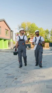 İki inşaat işçisi, yeni döşenmiş yeşil ve tuğlalı bir konağın yakınındaki asfalt bir alanda, projenin gidişatını tartışıyorlar.