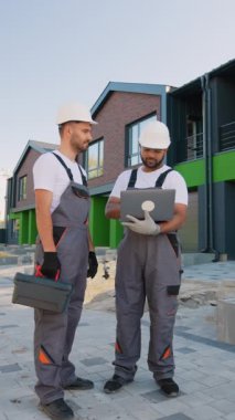 İki inşaat işçisi, inşaat başlıkları ve üniformalarıyla bir dizüstü bilgisayarda modern yeşil ve tuğlalı bir konağın yanında dikilirken planlarını gözden geçiriyorlar.