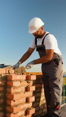 Hintli inşaat işçisi yeni bir ev için ustalıkla tuğla duvar örüyor, modern bir inşaat alanında güneşli gökyüzünün altına havan topu sıkıyor.