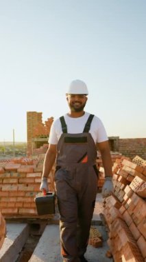 Güvenlik kaskı ve tulumlarıyla gülümseyen Hintli inşaat işçisi, güneşli bir inşaat alanındaki tuğla yığınlarının arasında güvenli bir şekilde yürüyor.