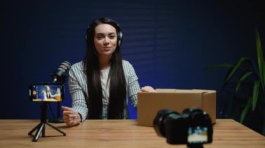 Stüdyoda video kaydı yapan bir kadın, akıllı telefon kamerasının önünde yeni bir kazakla kutusunu açan gülümseyen bir kadın.