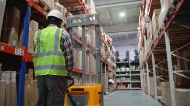 Sıkı bir şapka ve güvenlik yeleği giymiş Afro-Amerikan bir depo işçisi depo koridorunda yürüyor, iyi organize edilmiş bir tesiste forklift kullanıyor.