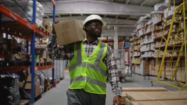Sıkı bir şapka ve güvenlik yeleği giymiş Afro-Amerikan teslimat işçisi bir depoya giriyor, organize depolama koridorlarında gezinirken omzunda bir kutu taşıyor.