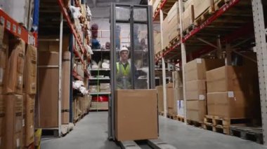Depo çalışanı sıkı bir şapka ve güvenlik yeleği ile malları depolama koridorlarında forklift kullanarak taşıyarak organize bir tesiste etkili operasyonlar sağlıyor.