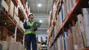 Güvenlik yeleği ve kasketi olan bir depo çalışanı, envanter verilerini bir dizüstü bilgisayardan kontrol ederek, etkin bir hisse yönetimi ve organizasyon sağlar.