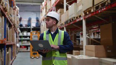 Güvenlik teçhizatı olan bir depo çalışanı dizüstü bilgisayar kullanarak envanteri kontrol ediyor. Verimli işlemler ve zamanında teslimat için doğru stok seviyelerinin sağlanması