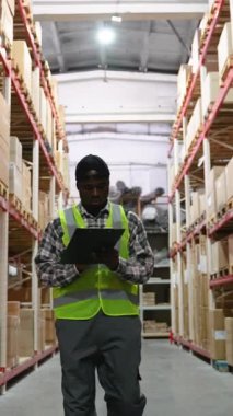 Güvenlik yeleği giyen Afrikalı Amerikalı işçiler, büyük bir depo koridorunda envanter kontrolü yapıyor, raflar dolusu kutu ve malla dolu.