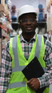 Afro-Amerikan lojistik işçisi, kasketli ve güvenlik yeleği ile depodaki koridorda yürüyor, rafları inceliyor ve envanter doğruluğunu sağlama alıyor.