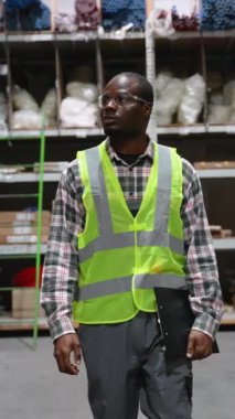 Afro-Amerikan lojistik işçisi, kasketli ve güvenlik yeleği ile depodaki koridorda yürüyor, rafları inceliyor ve envanter doğruluğunu sağlama alıyor.