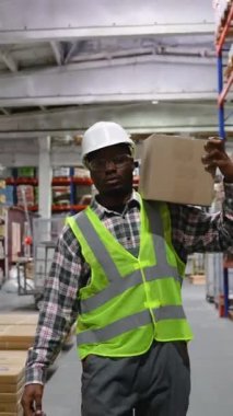 Güvenlik yeleği ve şapka takmış Afrikalı Amerikalı bir depo çalışanı bir depoya giriyor, envanter işlemlerini yönetirken bir kutu taşıyor.