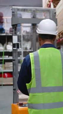 Güvenlik yeleği ve kasketli bir teslimatçı depoya giriyor, müşteriye teslimat için raflardan bir paket seçiyor.