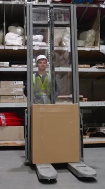 Depo çalışanı, organize koridorlardan büyük bir kutu mal taşımak için forklift kullanıyor. Teslimatı verimli teslimat operasyonları için hazırlıyor.