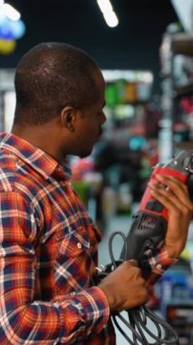 Ekose gömlekli Afro-Amerikalı bir adam, vitrindeki çeşitli alet ve aksesuarlarla çevrili bir hırdavatçıda alışveriş yaparken kırmızı elektrikli matkabı inceliyor.