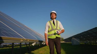 Kask ve güvenlik yeleği giyen bir mühendis, güneş enerjisi alanındaki operasyonları denetlerken proje planları tutuyor.