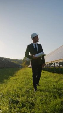 Yeşil bir alanda güneş panellerinin yanında yürüyen takım elbiseli bir mühendis yenilenebilir enerji, sürdürülebilirlik ve modern teknolojiyi temsil ediyor.