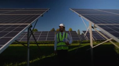Güvenlik yeleği ve miğferinde profesyonel mühendis berrak mavi gökyüzünün altındaki yenilenebilir enerji alanındaki güneş panellerini inceliyor.