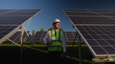 Güvenlik yeleği ve kask giyen profesyonel bir mühendis, güneşli bir günde yenilenebilir enerji alanındaki yeni kurulmuş güneş panellerini kontrol ederek kalite ve verimlilik sağlar.