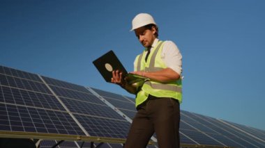 Bir sabit şapka ve güvenlik yeleği yatırımcısı güneş panellerinin yanında dururken dizüstü bilgisayardaki performans verilerini analiz eder.