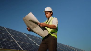 Sert şapka ve güvenlik yeleği giyen bir mühendis, güneş panellerinin yanında dururken projenin özellikleriyle açık bir gökyüzü altında aynı hizayı sağlayarak projeleri gözden geçirir.