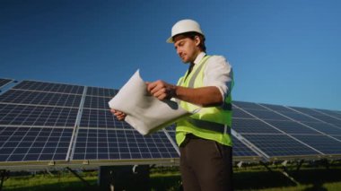 Kask ve güvenlik yeleği giyen bir mühendis yenilenebilir enerji alanındaki güneş panellerinin yanında dururken kurulum planlarını dikkatlice gözden geçirir.